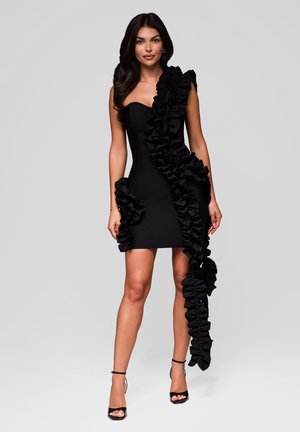 Kvinna klädd i en svart strapless mini-klänning med stora volanger som faller asymmetriskt, tillsammans med svarta klackar med ankeldetalj.