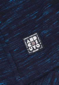 Marineblauwe gebreide stof met lichtblauwe spikkels, voorzien van een zwart logo-vlek met witte, grijze en blauwe tekst en een ontwerpelement.