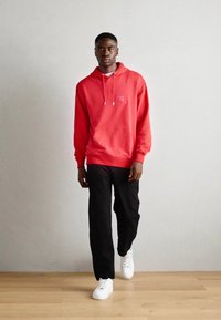 Rød hættetrøje med snore, sorte bukser og hvide sneakers. Har en frontlomme og et lille logo på brystet.