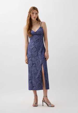 Robe longue sans manches de couleur bleu profond avec un motif abstrait, dotée d'une fente sur le côté et de fines bretelles. Accentuée par des sandales à talons hauts argentées.
