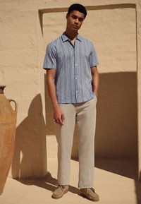 Chemise à manches courtes boutonnée de couleur bleu clair avec rayures verticales, associée à un pantalon en lin gris clair et des chaussures beige. Fond texturé.