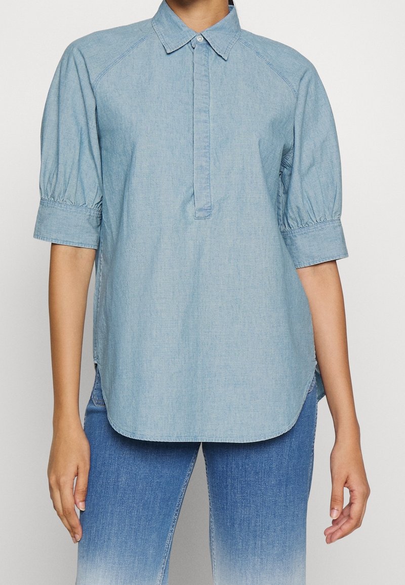 Chemise en denim bleu clair à manches courtes avec col et patte de boutons cachée, portée avec un jean ombré bleu sur un fond uni.