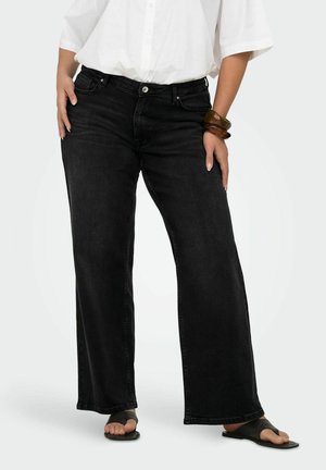 Osoba oblečená v čiernych džínsach s širokými nohavicami, bielej košeli na gombíky, čiernych sandáloch a hnedom náramku, stojaca s rukami pri bokoch.