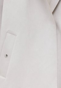 Manteau beige clair avec une poche à rabat dotée d'une fermeture par bouton-pression métallique, mettant en valeur un tissu lisse et une couture détaillée.