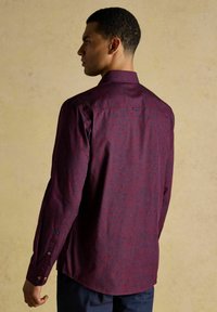 Vīrietis ar tumši sarkanu krekla ar garām piedurknēm, uz kura ir smalka ziedu raksts, stāv šķērsām pret vienkāršu beigu fona.