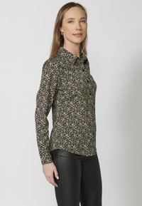 Camicia nera a maniche lunghe con stampa floreale in crema e giallo. Presenta un colletto, chiusura frontale con bottoni e un design aderente.