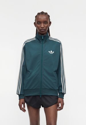 Person trägt eine tealfarbene Adidas Trainingsjacke mit Reißverschluss, weißen Streifen an den Ärmeln und Logo auf der Brust, kombiniert mit schwarzen Shorts, steht vor weißem Hintergrund.