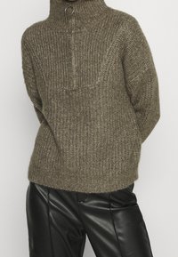 Pull tricoté vert olive avec col montant et demi-fermeture éclair. Tissu texturé et coupe décontractée. Associé à un pantalon en cuir noir.
