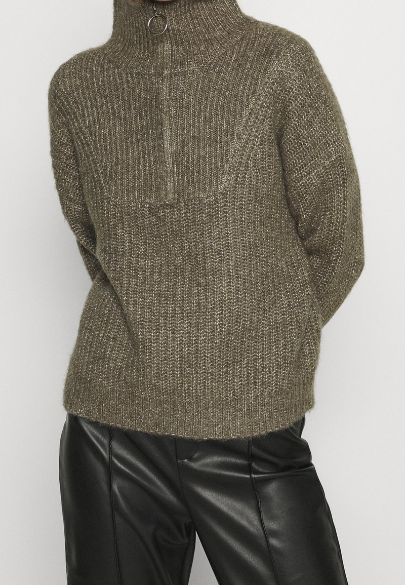 Pull tricoté vert olive avec col montant et demi-fermeture éclair. Tissu texturé et coupe décontractée. Associé à un pantalon en cuir noir.