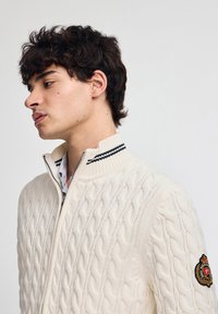 Vit zip-up kabelstickad tröja med hög krage och svarta randiga detaljer. Har en logopatch på vänstra ärmen.