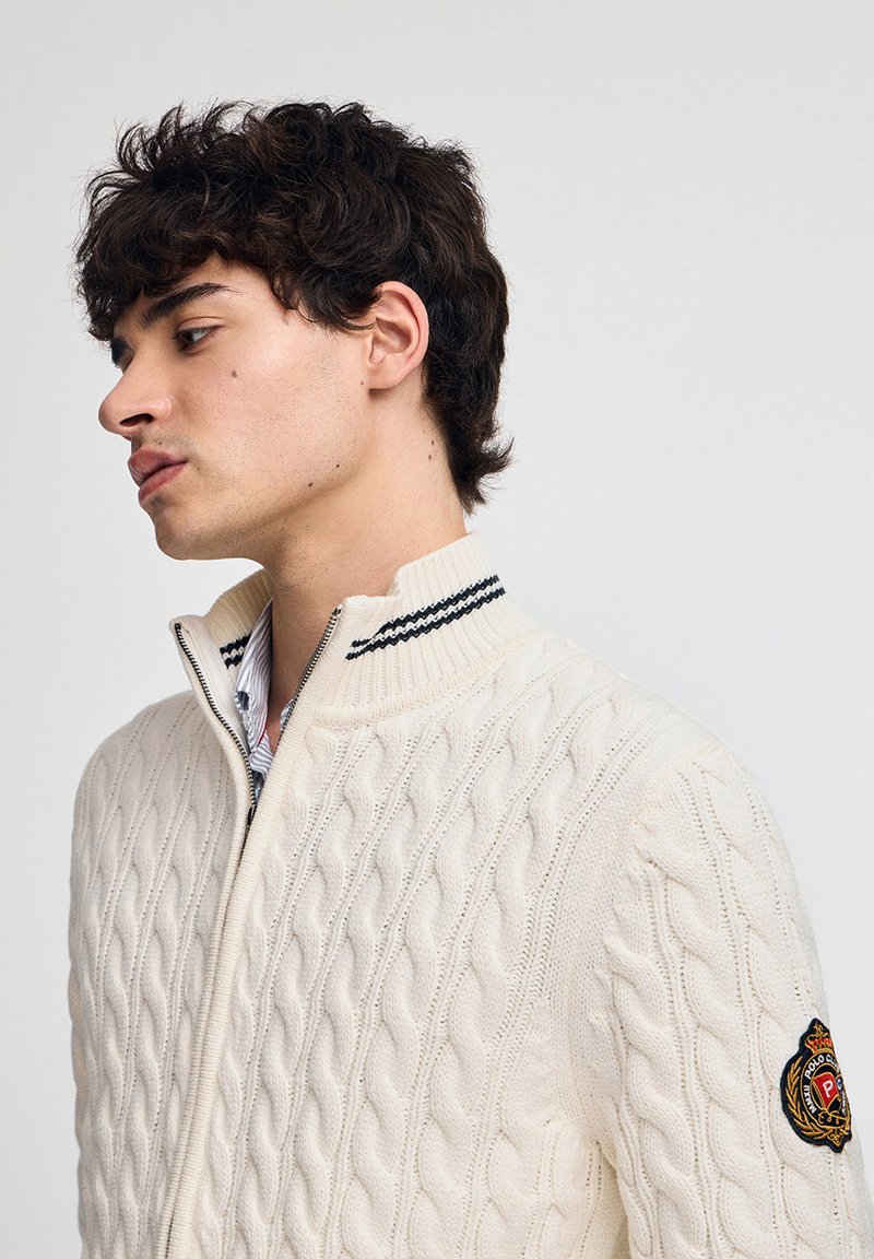 Vit zip-up kabelstickad tröja med hög krage och svarta randiga detaljer. Har en logopatch på vänstra ärmen.