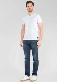 T-shirt en coton blanc à col en V associé à un jean slim bleu foncé et des baskets blanches, avec une petite étiquette de marque sur l'ourlet de la chemise.