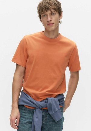 Mand med kort brunt hår iført en orange kortærmet T-shirt, blå jeans og en blå sweatshirt bundet omkring livet.