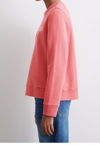 Coral sweatshirt, crew neck, soft fabric, relaxed fit, ribbed cuffs and hem, paired with blue jeans, minimalistic logo on the chest.