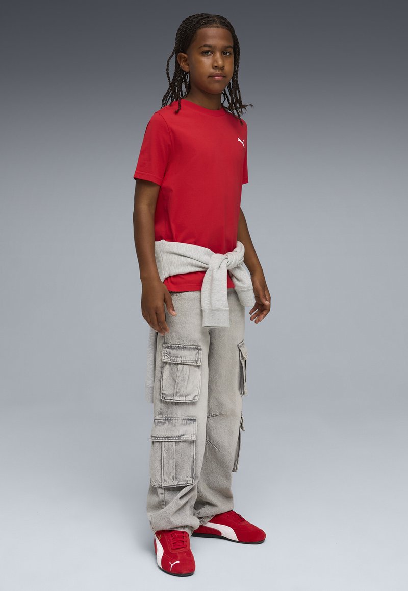 Παιδί με πλεξούδες που φοράει κόκκινο t-shirt Puma, ανοιχτόχρωμο γκρι παντελόνι cargo, δεμένο γκρι φούτερ και κόκκινα αθλητικά παπούτσια Puma, στέκεται μπροστά σε γκρι φόντο.