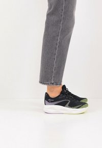 Chaussures de sport noires avec une semelle blanche et vert néon, présentant des rayures diagonales sur le côté et une tige en tissu texturé.