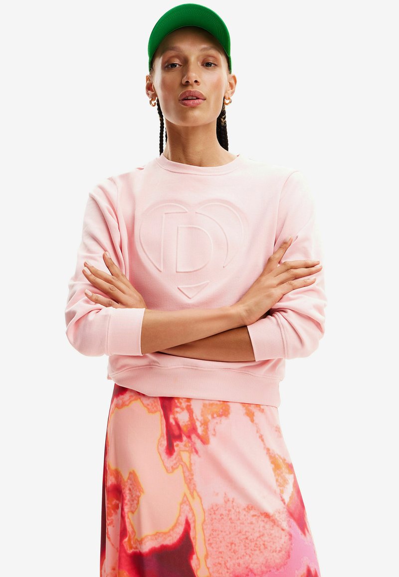 Sudadera rosa con logo en relieve, cuello redondo, puños acanalados, combinada con una falda de patrón abstracto colorido y una gorra verde.
