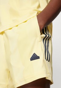 Žlté atletické šortky vyrobené z mäkkej látky, s čiernym logom Adidas a tromi vertikálnymi čiernymi pruhmi po stranách.