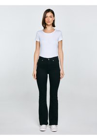 Sieviete stāv un skatās uz priekšu, valkājot baltu īsroku kreklu, melnas augsta vidukļa taisnā bikšu bikses ar pieguļošiem galiem un baltas sporta kurpes uz vienkārša fona.