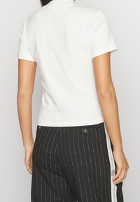 Bílý krátký rukáv, přiléhavý top s vysokým límcem. Hladká textura, minimalistický design. V kombinaci s černými pruhovanými kalhotami s bílými bočními pruhy.