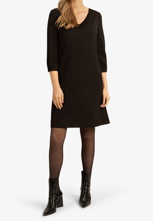 Robe noire avec un décolleté en V, des manches trois-quarts et une coupe loose qui arrive au-dessus du genou. Associée à des bottines noires à talons pointus.