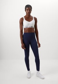 Hvid Nike sports BH med sort logo, parret med højtaljede navy leggings og hvide sneakers. Glat tekstur, tætsiddende design.