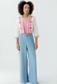 Witte cropped jas met roze bloemenborduurwerk, gecombineerd met een roze top en lichtblauwe wijde jeans. Horizontale naad en knoopdetails zichtbaar.