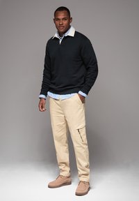 Dunkler Pullover mit einem weißen Kragen, hellblauem Hemd darunter, beigen Cargo-Hosen und beigen Stiefeln. Die Taschen der Hose sind sichtbar.