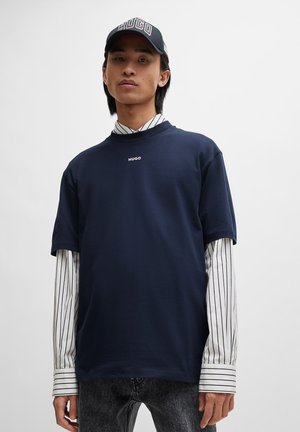 T-shirt en coton marine à col rond et manches courtes, arborant un petit logo "HUGO" blanc. Superposé avec une chemise à rayures blanches en dessous.