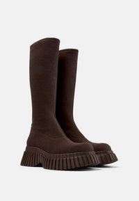 Botas altas de tela marrón con suelas de goma gruesas y estriadas y puntas redondeadas, colocadas una al lado de la otra sobre un fondo liso.