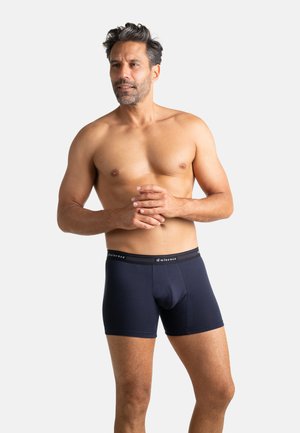 Homme torse nu aux cheveux courts et foncés portant un boxer Eminence bleu foncé, debout les mains jointes sur un fond blanc uni.