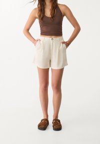 Débardeur ajusté marron avec un short crème présentant des poches latérales, une fermeture à bouton et un tissu texturé. Des sandales marron à lanières complètent la tenue.