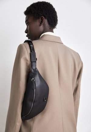 Person iført en beige blazer med en sort læder skuldertaske mod en ensartet lys baggrund, vist bagfra og fra siden.