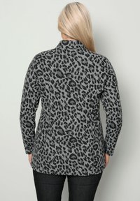 Donna con lunghi capelli biondi, che indossa un top a maniche lunghe grigio con stampa leopardata e pantaloni neri, voltata di spalle alla macchina fotografica.