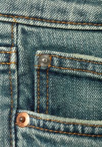 Detailný záber na džínsy z denimu, ktorý zobrazuje prednú kapsu, oranžové šitie a kovové nitovanie na modrom, vyblednutom textilnom povrchu.