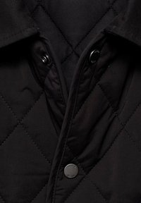 Chaqueta negra acolchada con textura suave, primer plano de un cierre de botón y un cuello con un acabado suave. Patrón geométrico de diamantes visible.