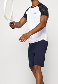 Valkoinen urheiluvaate, jossa on teksturoidut mustat lyhyet hihnat, yhdistettyinä tummansinisiin shortseihin. Henkilö pitää tennismailaa, valmiina lyömään palloa.