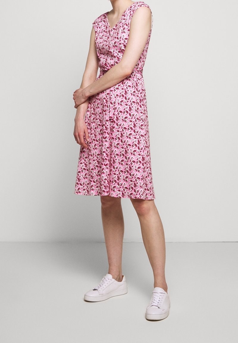 Květinové růžové šaty s krátkými rukávy, výstřihem do V, zúženým pasem a splývavou sukní. V kombinaci s bílými teniskami pro neformální vzhled.