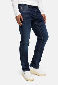 Dunkelblaue Denim-Jeans mit geradem Schnitt, dezenter Ausbleichung, kupferfarbenen Nähten und klassischem Fünf-Taschen-Design, getragen mit weißen Sneakers.