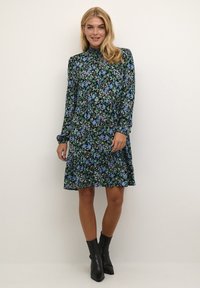 Langärmliges schwarzes Kleid mit einem floralem Muster in Blau und Grün. Es hat einen hohen Kragen und einen ausgestellten Rock; kombiniert mit schwarzen Stiefeletten.