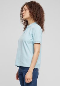 Šviesiai mėlynas polo marškinėlis trumpomis rankovėmis, su klasikine apykakle ir laisvesniu modeliu, derinamas su mėlynomis džinsinėmis kelnėmis ir garbanota, laisvai nusagstyta šukuosena.