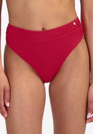 Slip bikini rosso testurizzato a vita alta indossato da donna in piedi, che mostra torso, fianchi e parte superiore delle cosce su sfondo bianco.
