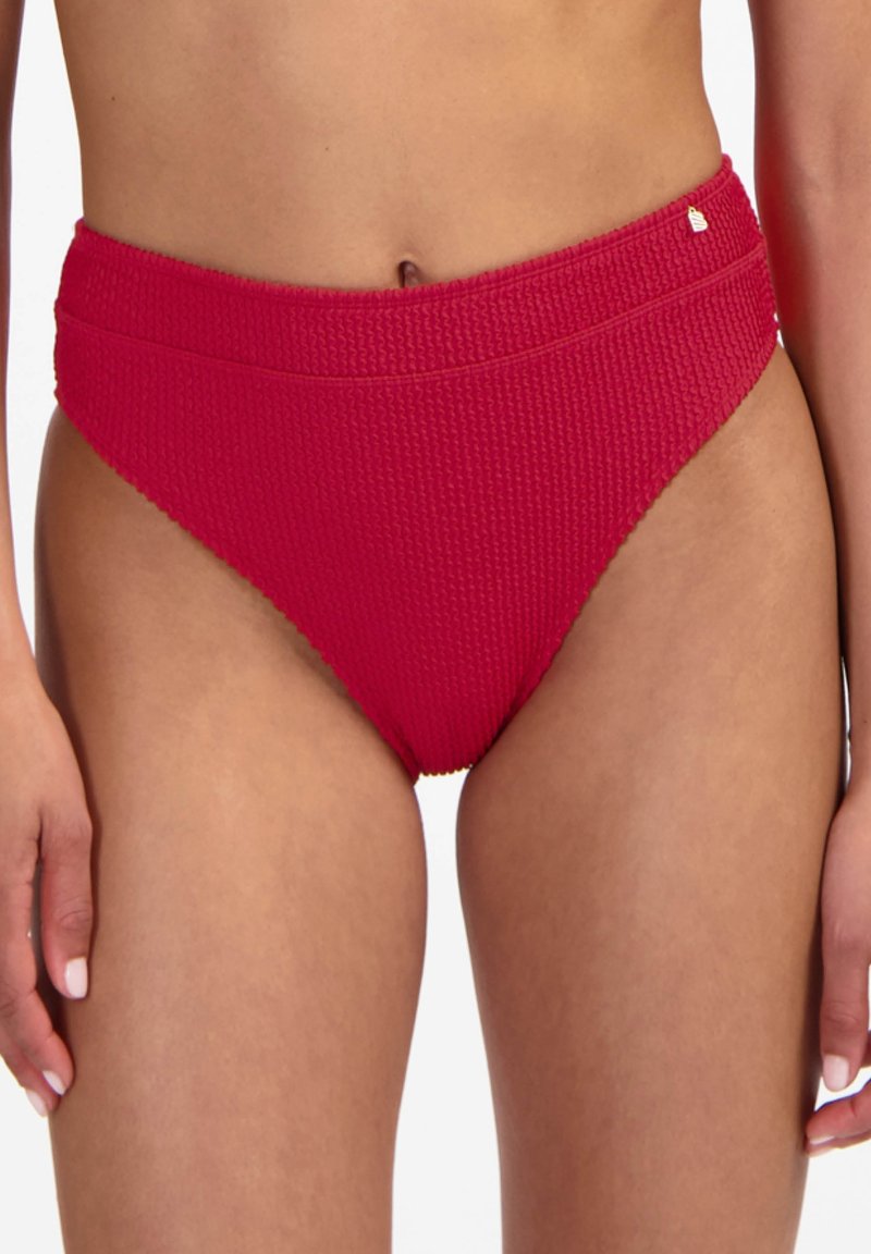 Bas de bikini rouge texturé taille haute porté par une femme debout, montrant le torse, les hanches et le haut des cuisses sur un fond blanc.