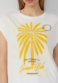 Witte T-shirt met een grote gele palmboom graphic, tekst "Vergeet niet om lid te worden van onze Zomerclub," met een zachte textuur.