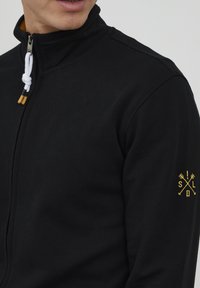 Schwarze Zip-Jacke mit hohem Kragen, weißem Kordelzug und Holzakzenten. Mit gelbem gesticktem Logo auf dem linken Ärmel.
