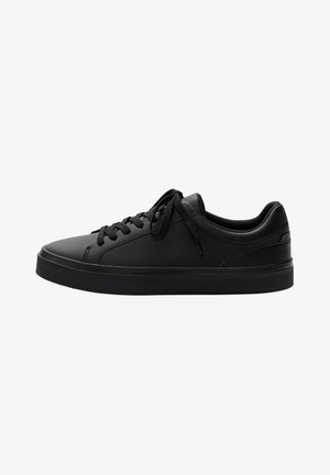 Zapatilla negra de caña baja con cierre de cordones delantero, parte superior de cuero liso, cuello acolchado y suela plana de goma, mostrada desde el lado exterior.