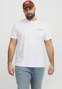 Weißes Baumwoll-T-Shirt mit kurzen Ärmeln, das auf der linken Brustseite ein kleines Logo in dunkler Schrift zeigt. Getragen mit hellblauen Jeans.