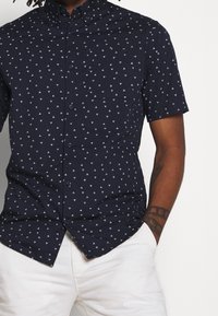 Camicia a maniche corte navy con un motivo triangolare bianco, design con bottoni, colletto piccolo e taschino sul petto.
