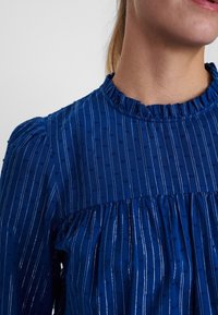 Blusa a righe blu con scollo arricciato e tessuto testurizzato. Piccole macchie sollevate creano un motivo in tutto il capo. Design svasato e arricciato all'orlo.
