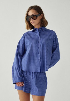 Frau trägt ein übergroßes blau kariertes Hemd mit Knöpfen und passende Shorts sowie große schwarze Sonnenbrillen vor einem einfarbigen Hintergrund.